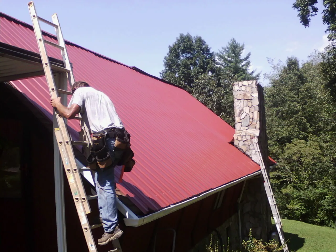 Roofer on a ladder beside a red metal roof and stone chimney — Soffit & Fascia Repair by Roofing Brothers in Montreat